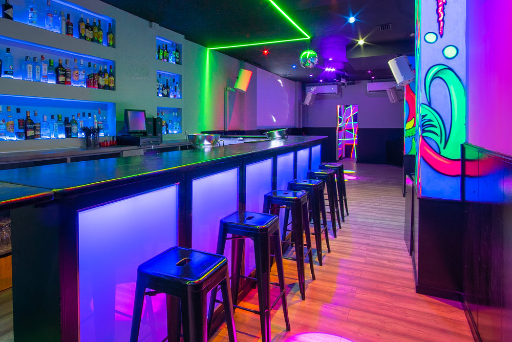 Sala Purple en Barcelona, barra larga retroiluminada en morado con taburetes altos, neones de colores y mural fluorescente