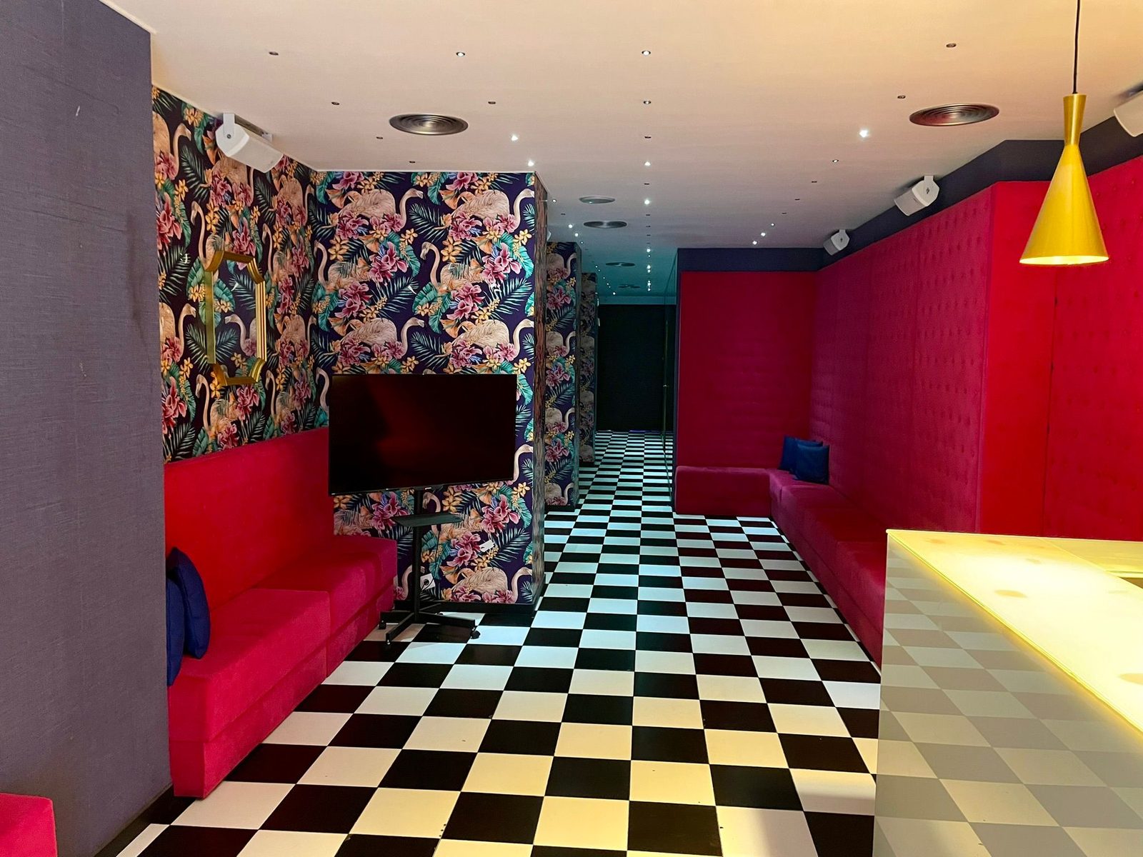Sala L. Silver en Barcelona con papel pintado tropical de flamencos, suelo de damero y sofás rojos junto a la barra iluminada
